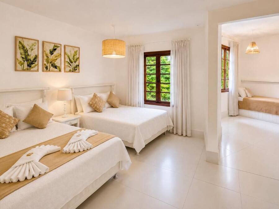 Una habitación de hotel con dos camas, una ventana con cortinas, una lámpara fijada a la pared y una puerta que da a otra habitación.
