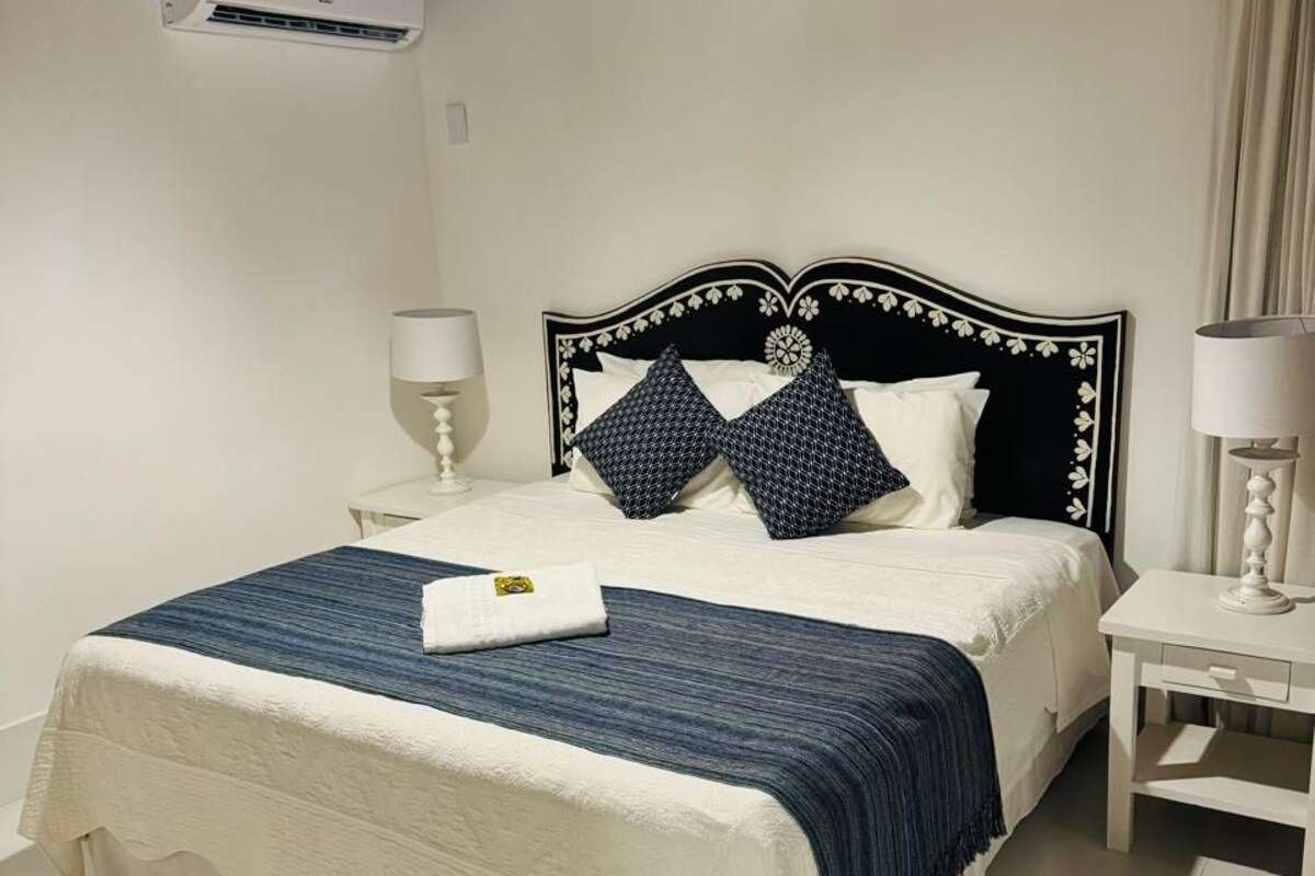 Uma cama bem arrumada com colcha branca e azul, duas almofadas iguais e um painel decorativo. Há uma mesa de cabeceira com abajur em cada lado da cama.