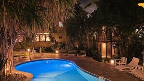 Ein Pool mit gewölbter Kante, umgeben von Liegestühlen und Palmen, mit einem Gebäude im Hintergrund.
