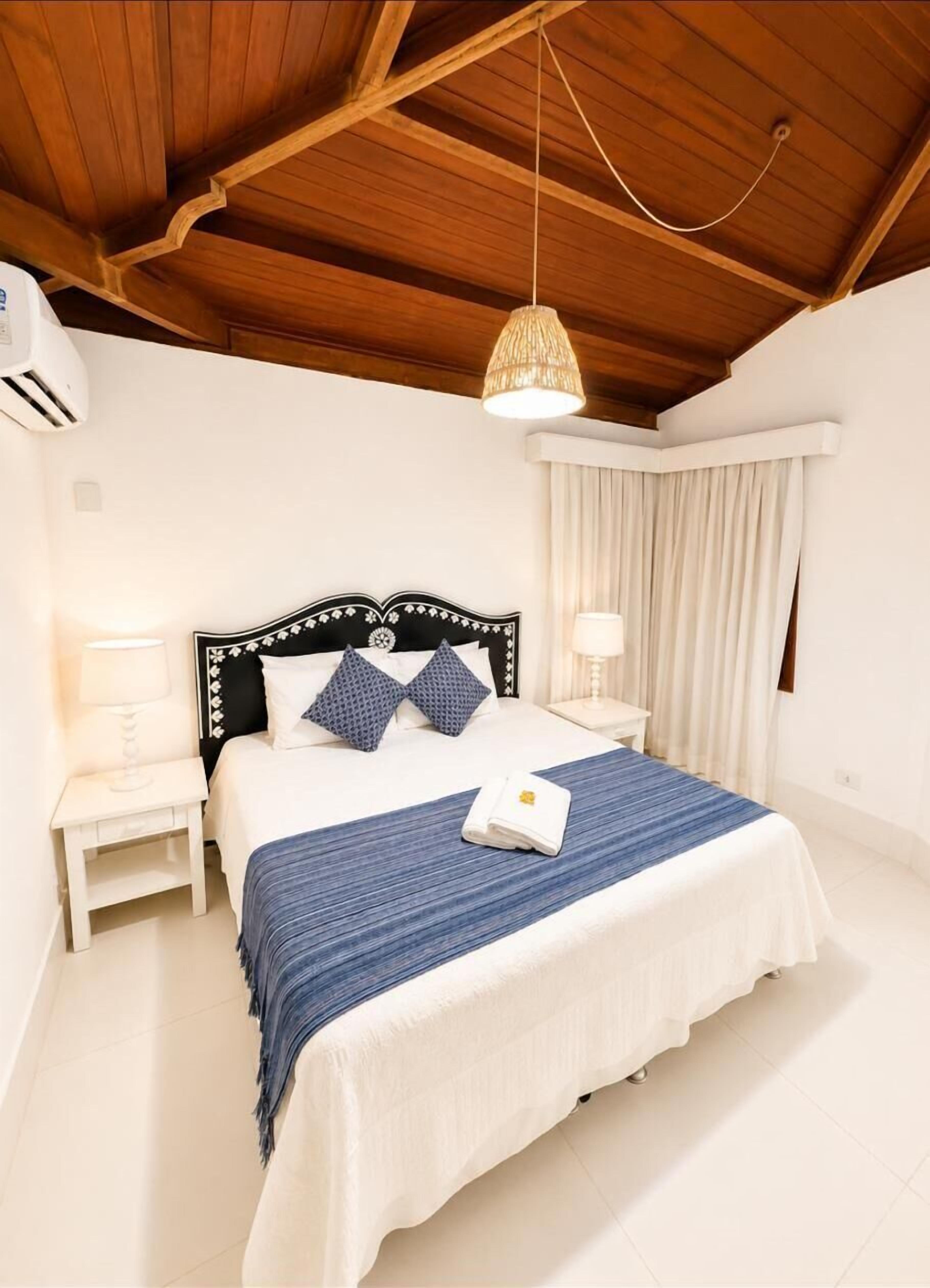 Een slaapkamer met een houten plafond, een bed met een blauwe spreideken, twee bedlampjes en een hangende lamp.