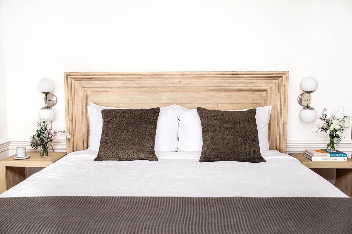 A neatly made bed with two dark pillows, a wooden headboard, white bedding, and a bedside table with a lamp and a vase of flowers.