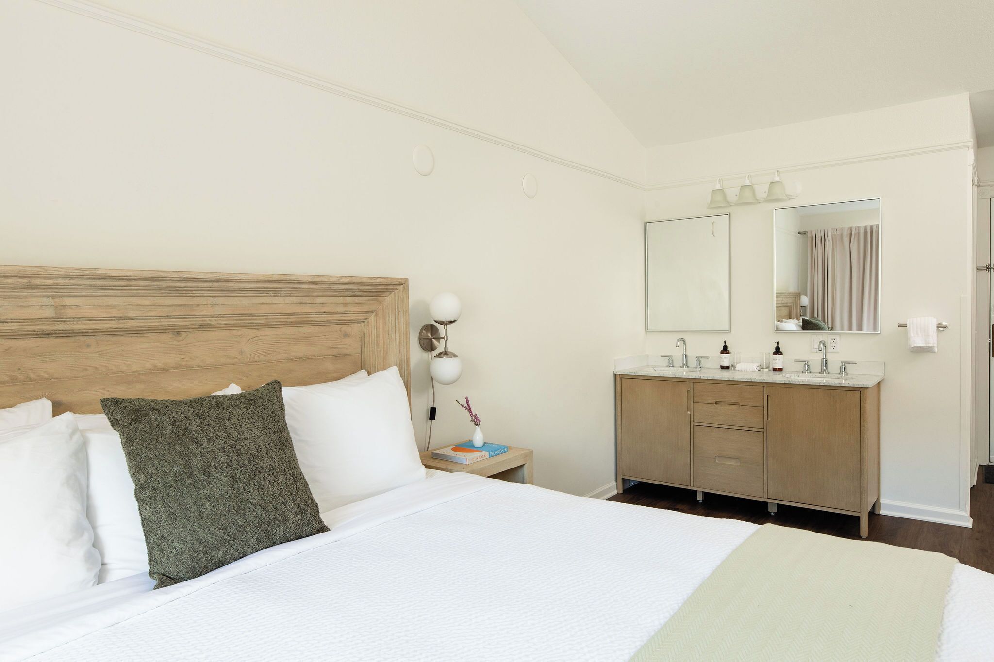 Une chambre d’hôtel avec une tête de lit en bois, un grand lit avec une literie blanche, une table de chevet avec une lampe et une salle de bain avec un meuble-lavabo en bois et un miroir.
