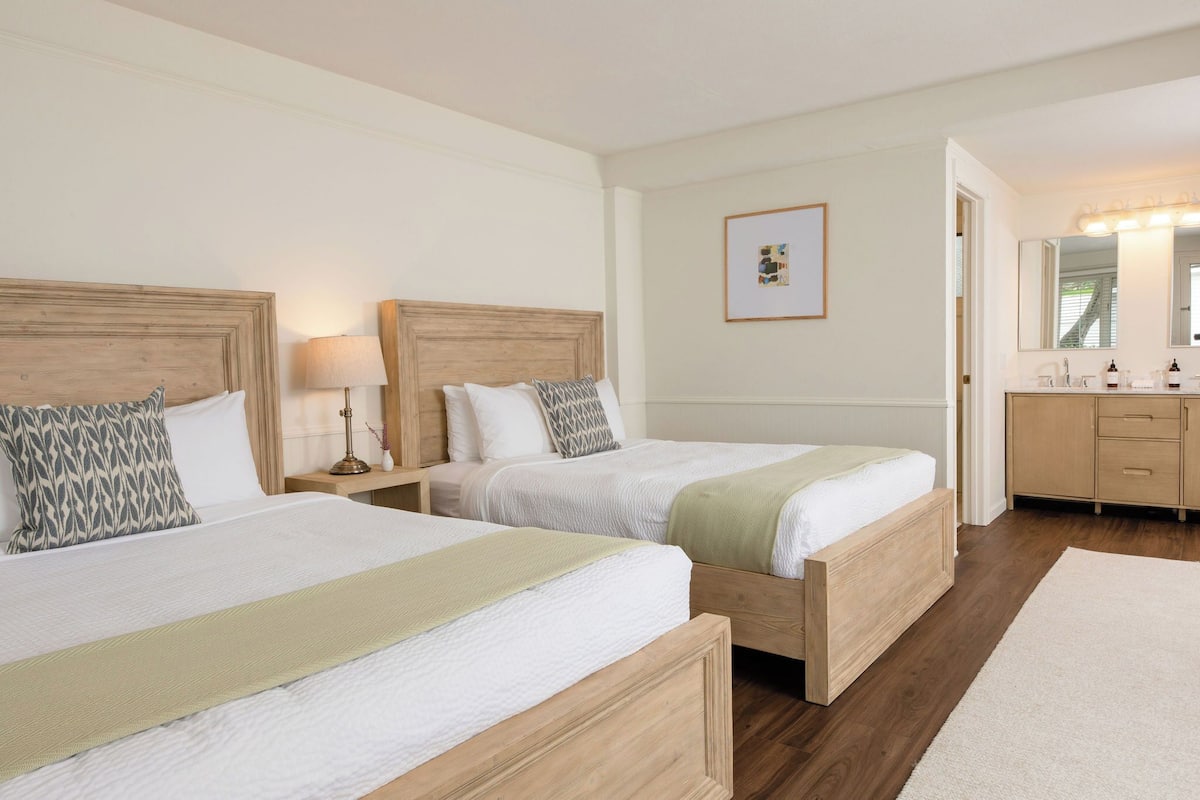 A hotel room with two beds, a wooden headboard, a bedside table with a lamp, a framed picture on the wall, and a bathroom with a vanity and mirror.