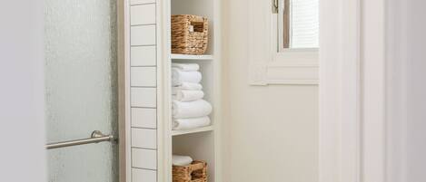 A bathroom with a white tiled wall, a glass shower door, and a white storage cabinet with wicker baskets and folded towels.