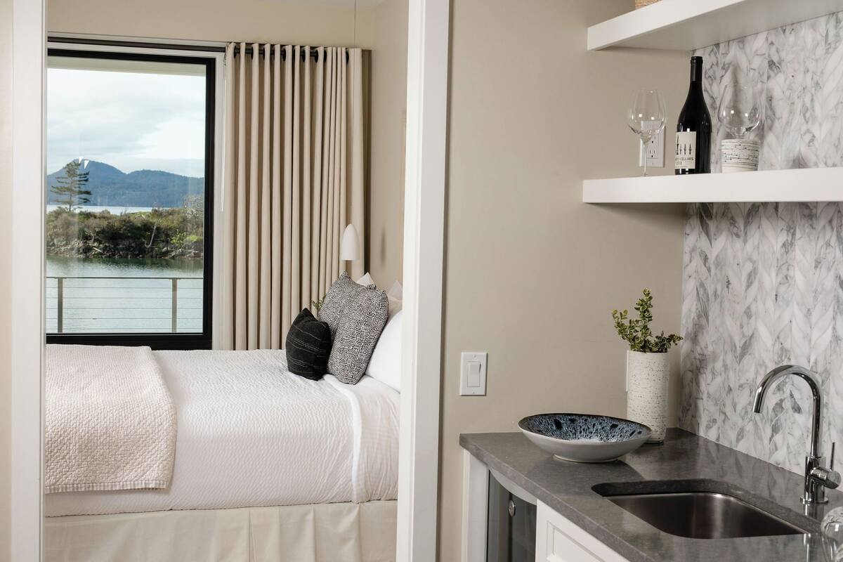 A bedroom with a view of a lake and mountains, a bed with white bedding, a bedside table with a lamp, and a bathroom with a marble backsplash and sink.