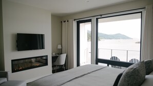 Een hotelkamer met een bed, een aan de muur bevestigde televisie, een bureau met stoel en een groot raam met uitzicht op zee en bergen.