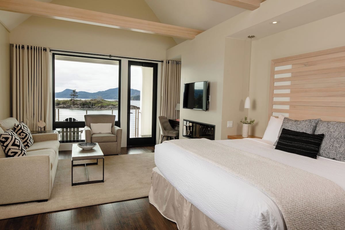 A hotel room with a large bed, a sofa, a chair, a TV, and a view of a lake and mountains.