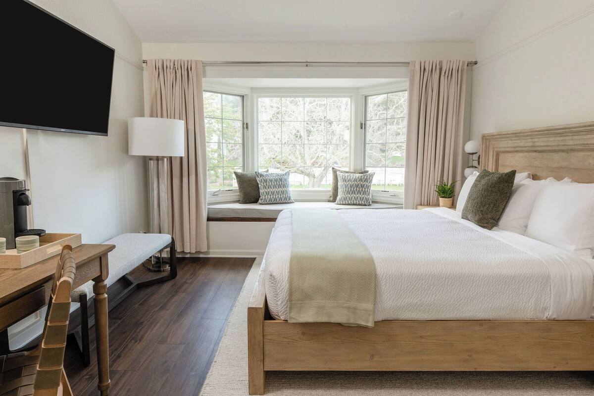 A bedroom with a large bed, a desk with a chair, a television, and a window with curtains.