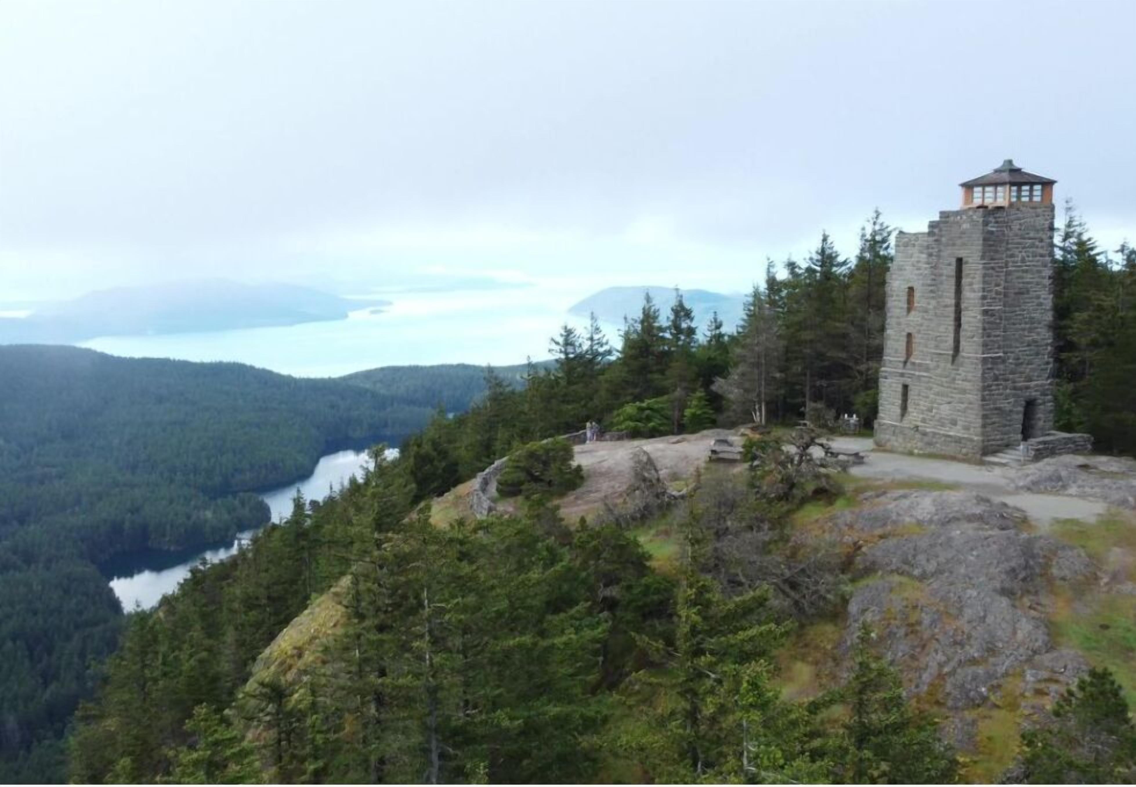 A stone tower with a lookout point, surrounded by dense forest and a lake in the background.