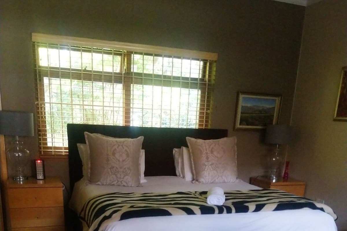 A neatly made bed with a zebra-patterned blanket, a wooden nightstand with a lamp, and a window with blinds.