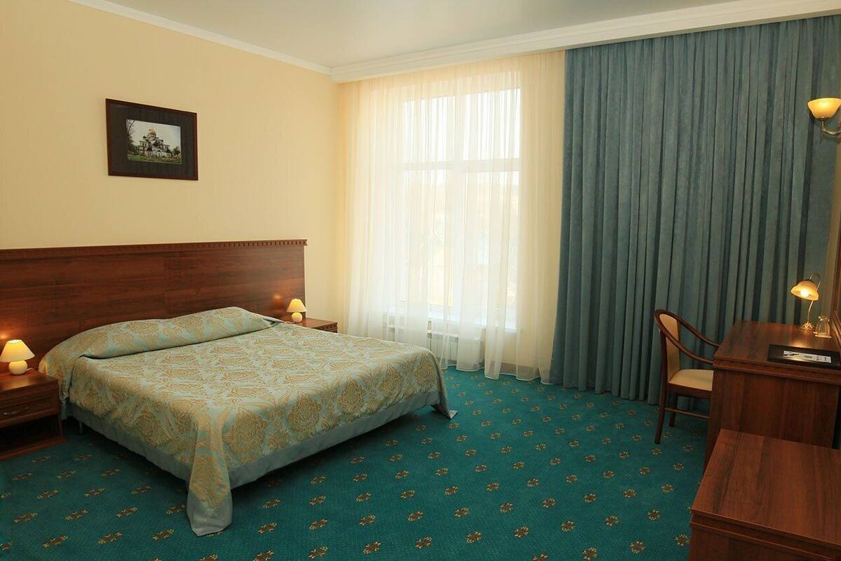 A hotel room with a bed, a desk, a chair, a nightstand, a lamp, a picture on the wall, and a window with curtains.