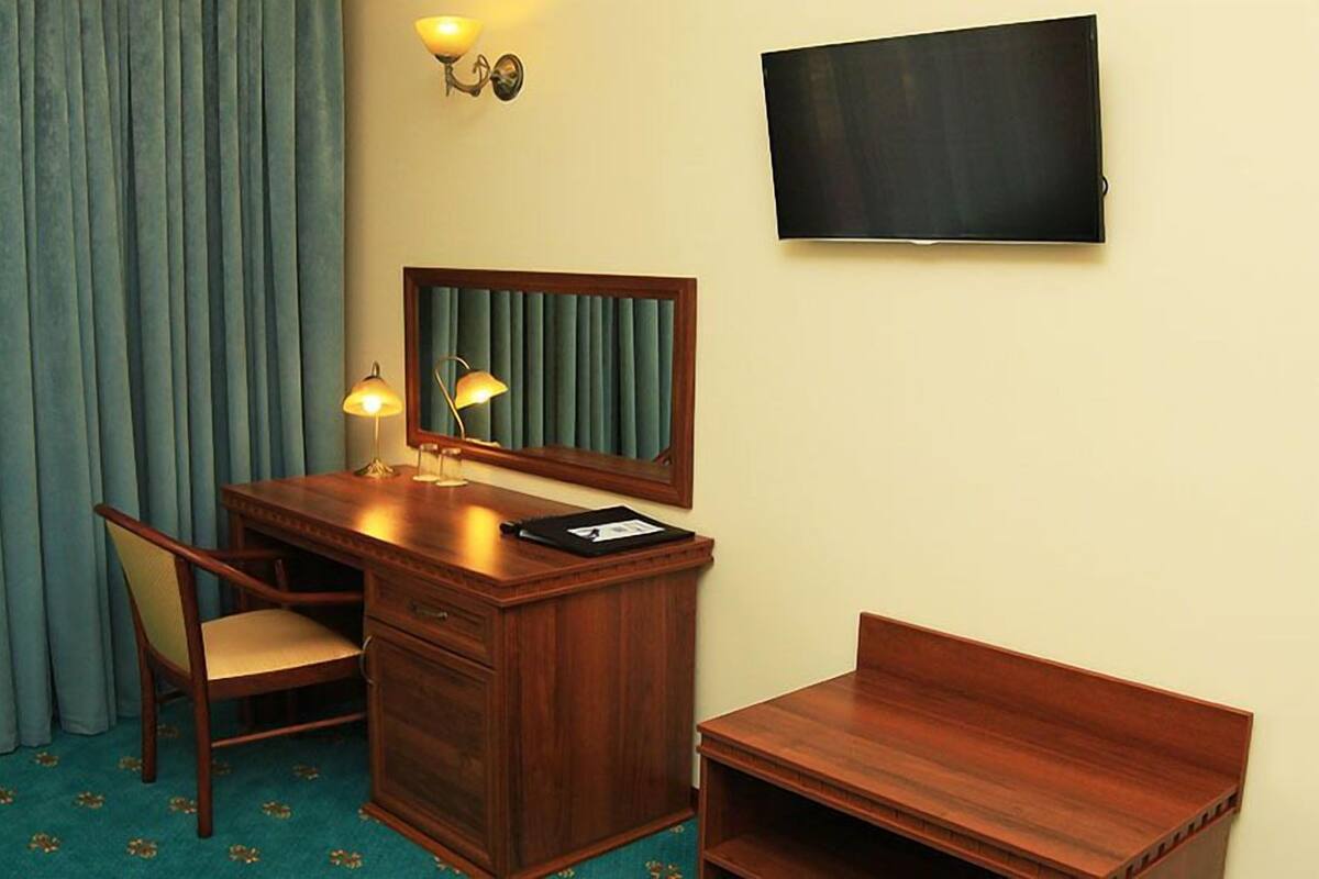 A hotel room with a wooden desk, a chair, a small bench, a television mounted on the wall, and a window with curtains.