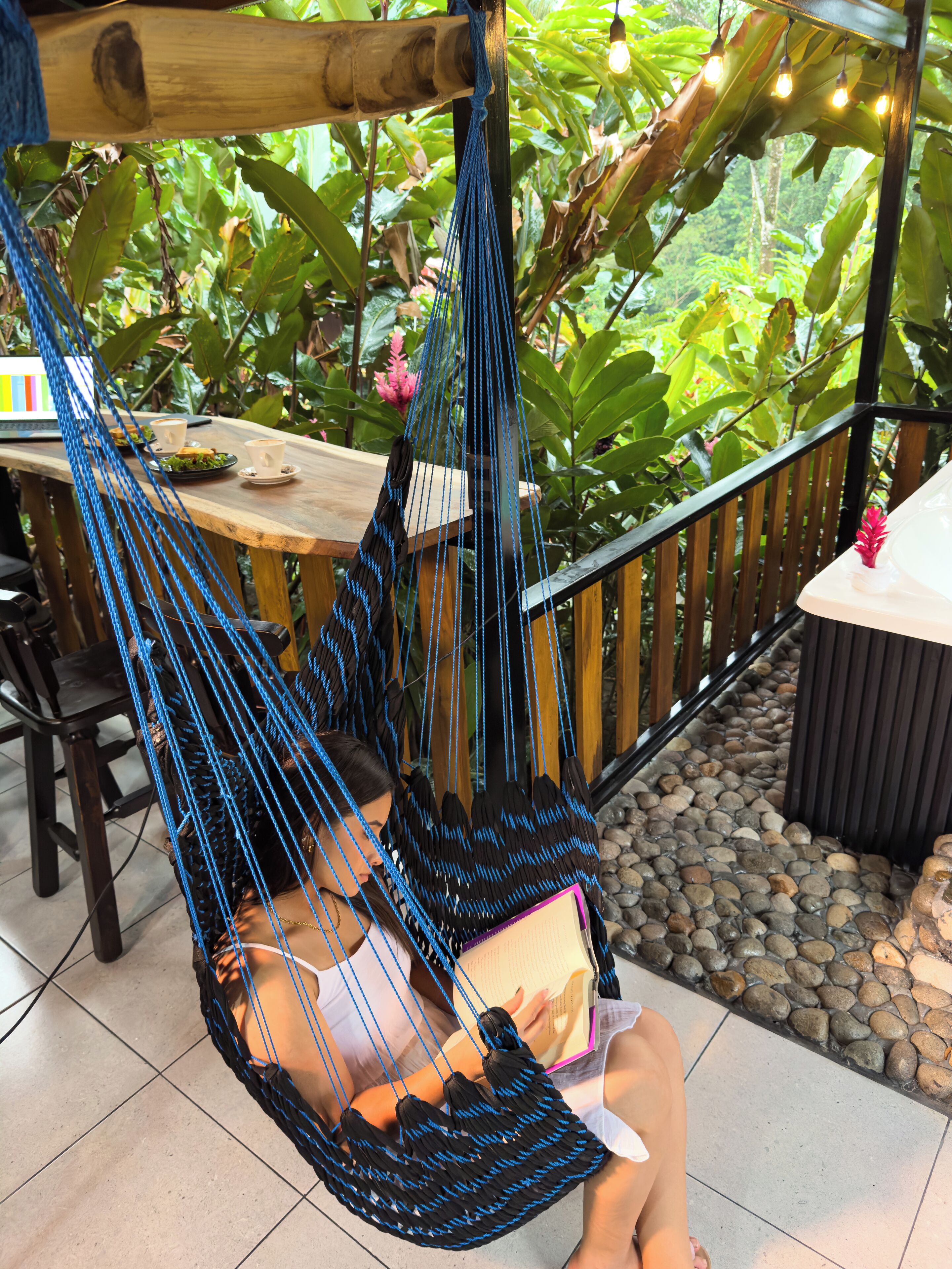 A hammock chair suspended from a wooden beam, a table with a laptop and cups, and a garden with plants and a stone pathway.