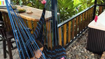 A hammock chair suspended from a wooden beam, a table with a laptop and cups, and a garden with plants and a stone pathway.