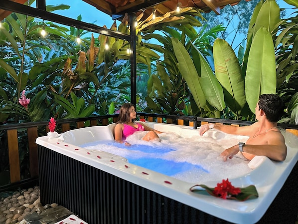 A hot tub with two people, surrounded by lush greenery and string lights.