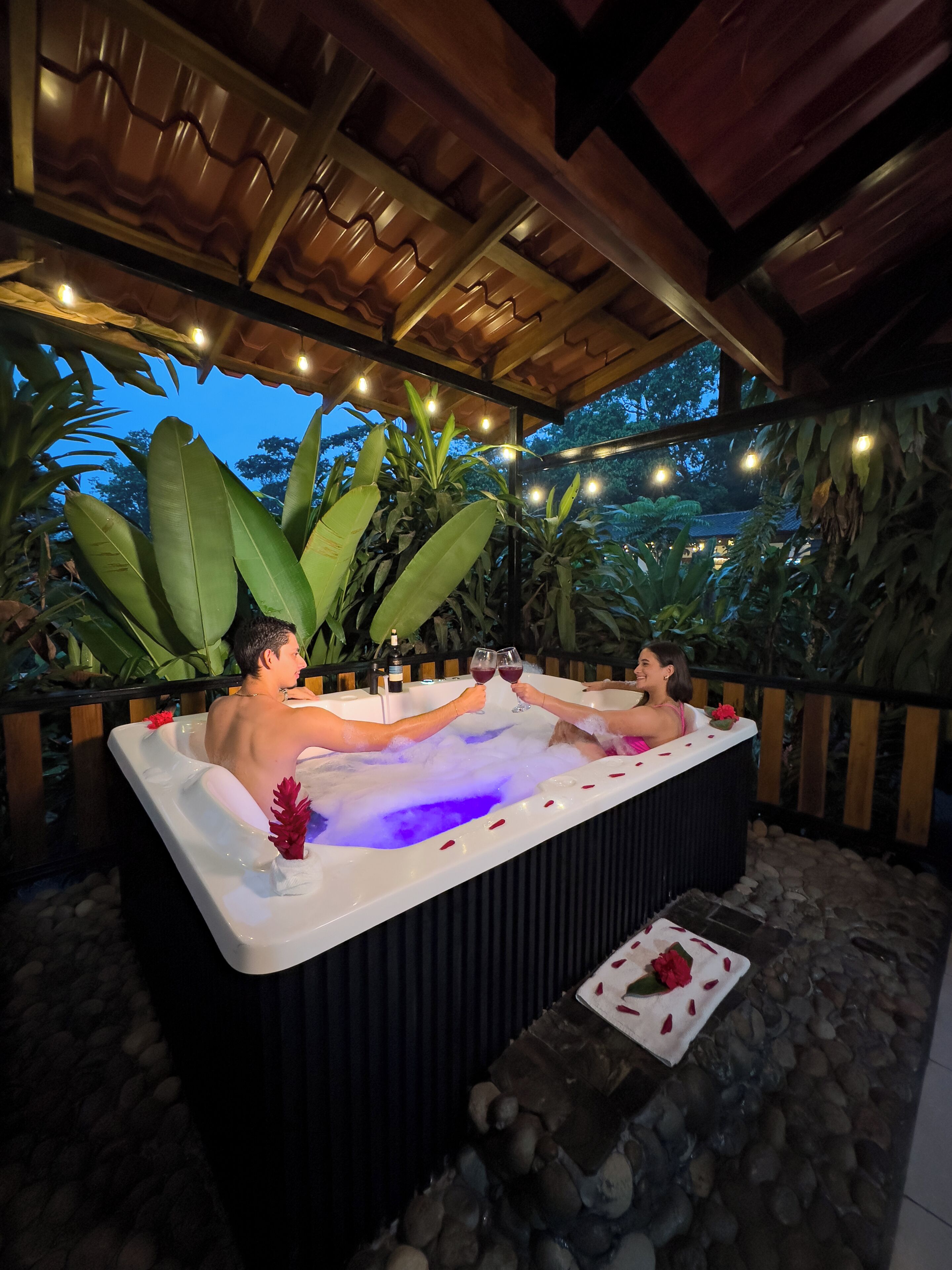 A hot tub with two people toasting with wine glasses, surrounded by tropical plants and string lights.