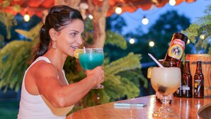 Une personne tenant un verre bleu dans un bar en plein air, avec des meubles en bois et une bouteille de spiritueux posée sur le comptoir.