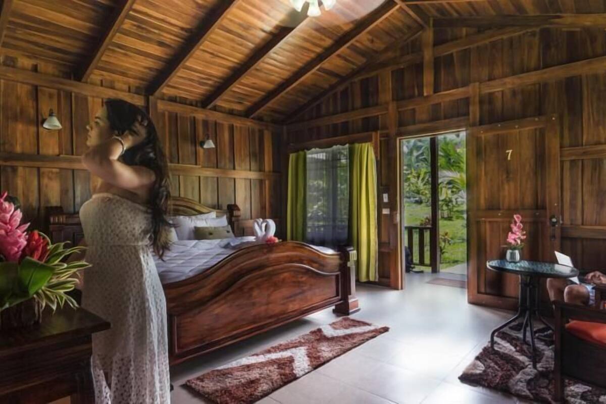 A wooden cabin with a large bed, a dining area, and a view of greenery outside.