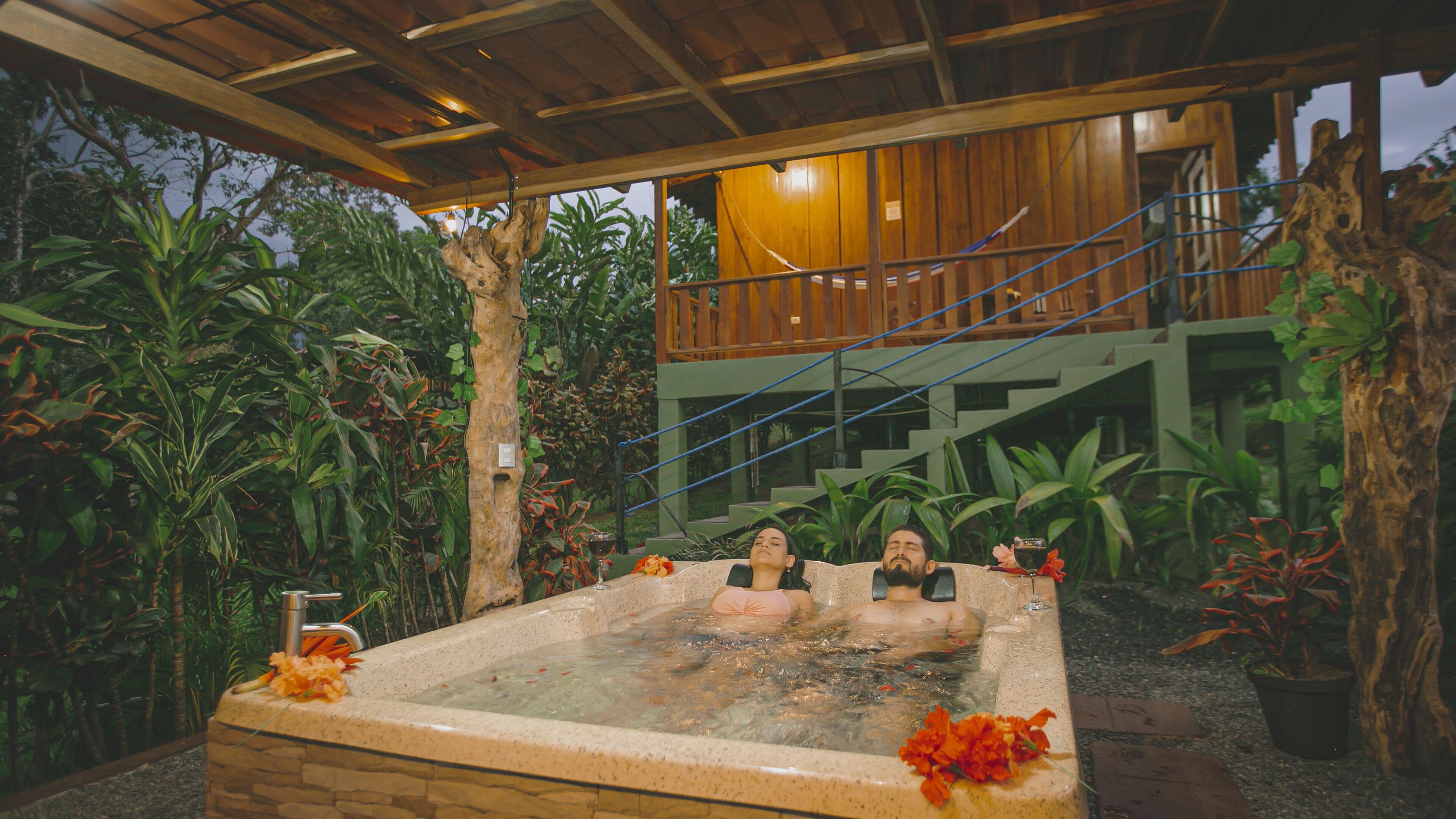 A hot tub with two people, surrounded by tropical plants and a wooden deck.