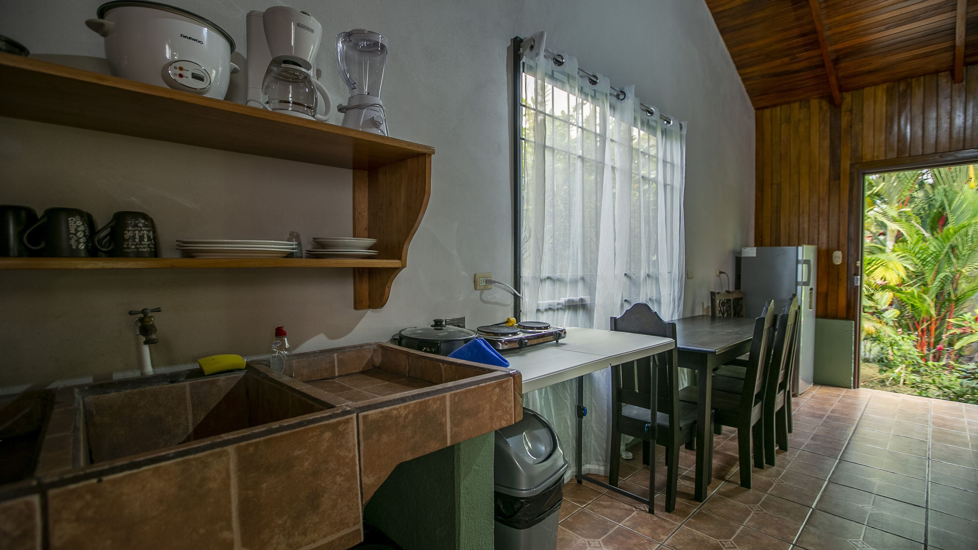 A kitchen with a wooden countertop, a sink, a coffee maker, a blender, and a window with curtains.