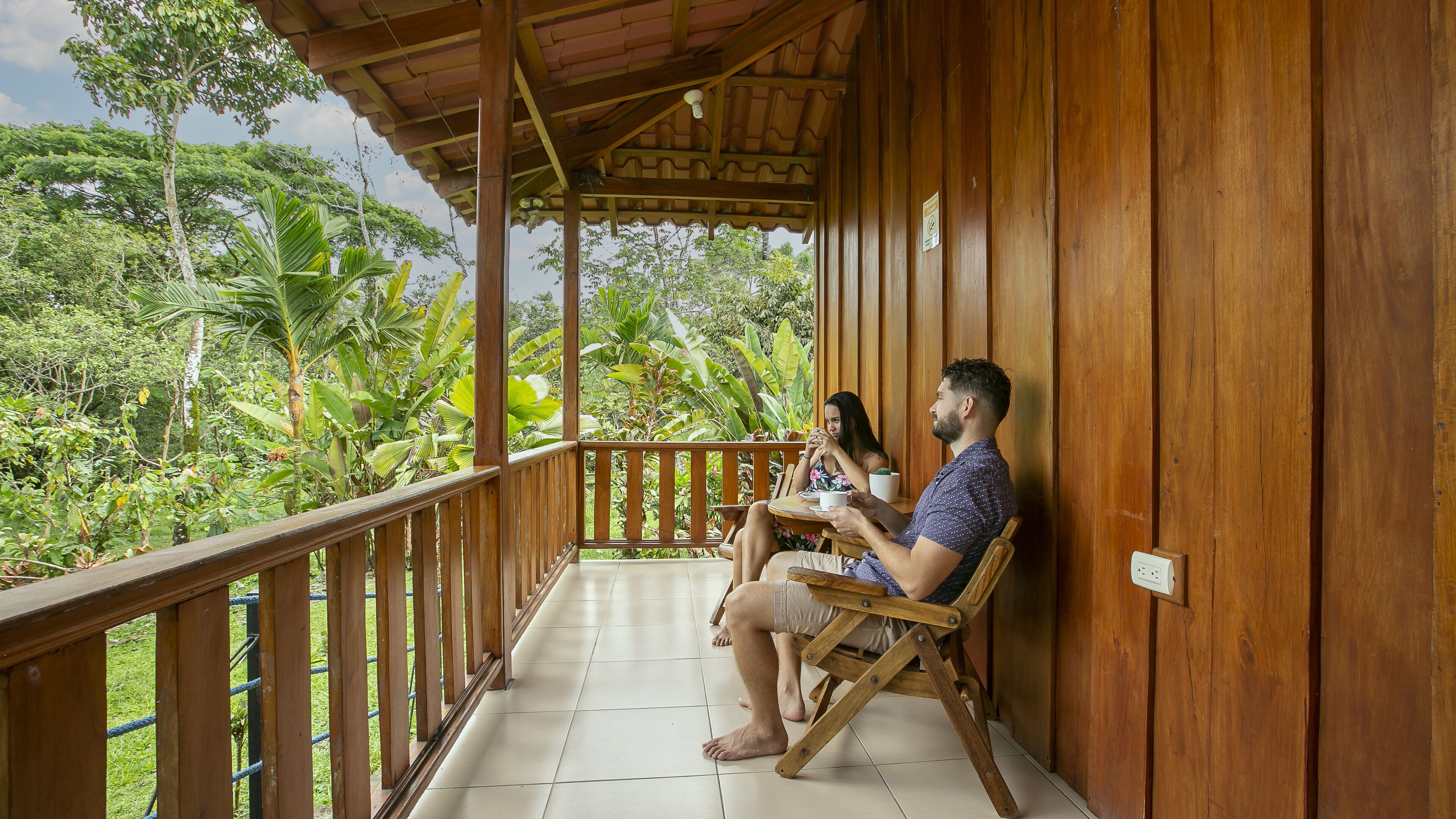 A wooden balcony with a tiled floor, overlooking a lush garden with various plants and trees.