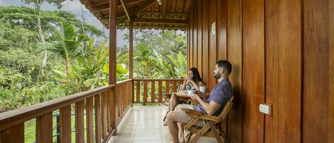 A wooden balcony with a tiled floor, overlooking a lush garden with various plants and trees.