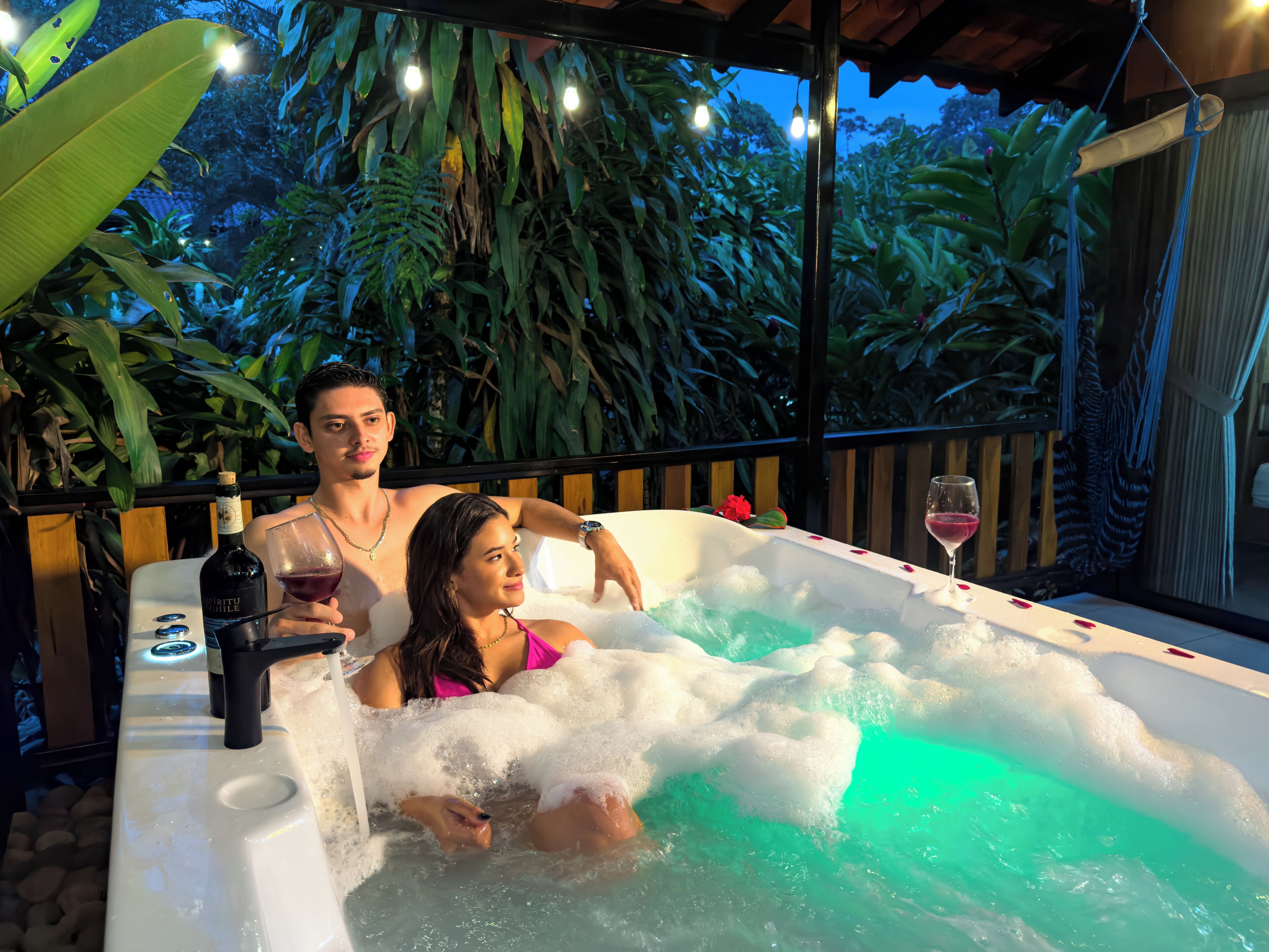 A couple in a hot tub with a bottle of liquor and a glass of wine, surrounded by lush greenery and string lights.