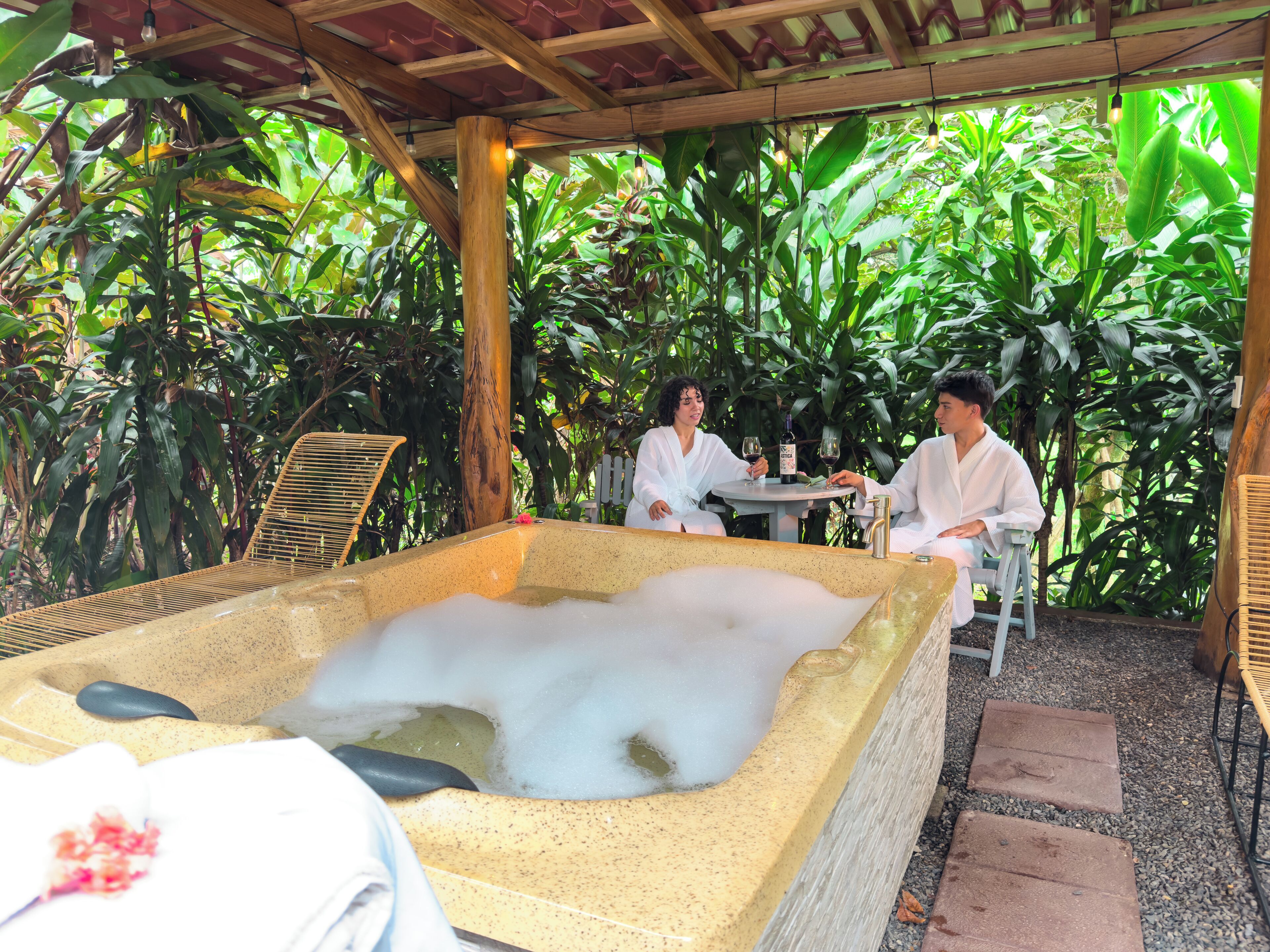 A spa area with a hot tub, two people seated at a table, surrounded by lush greenery.
