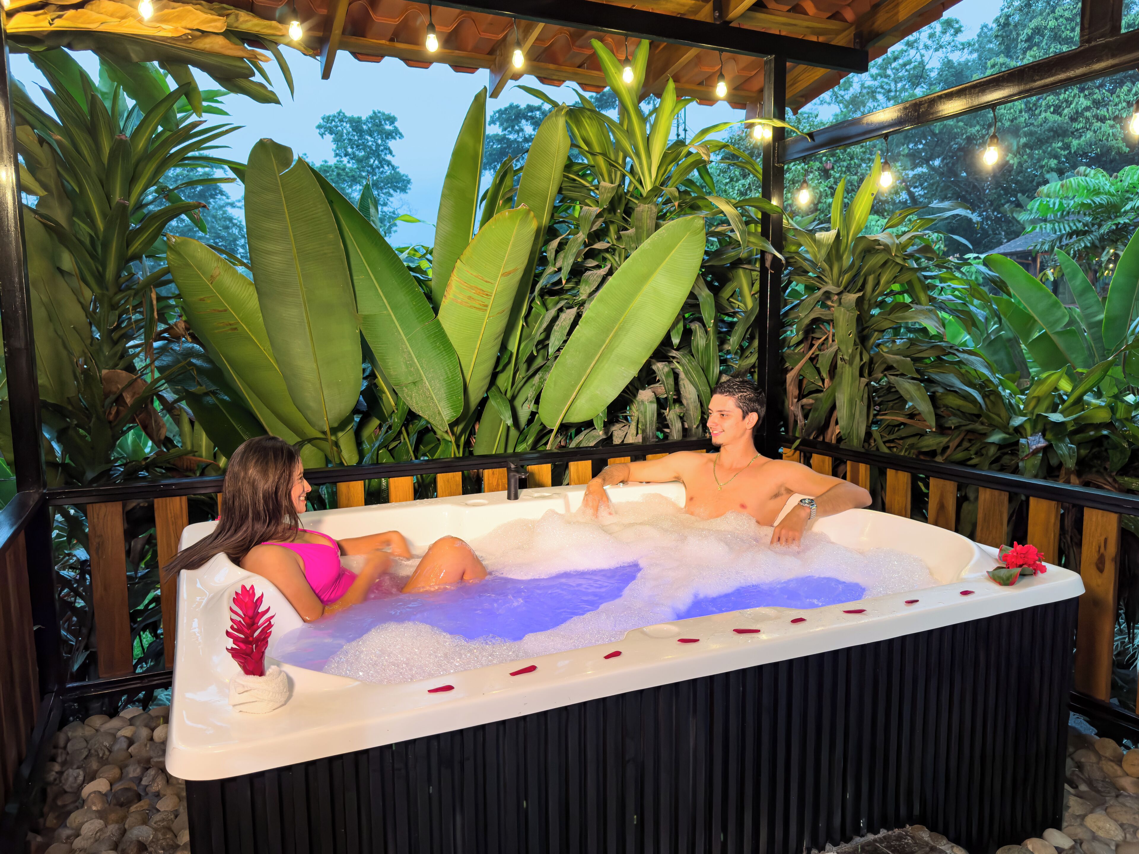 A hot tub with two people, surrounded by tropical plants and string lights.
