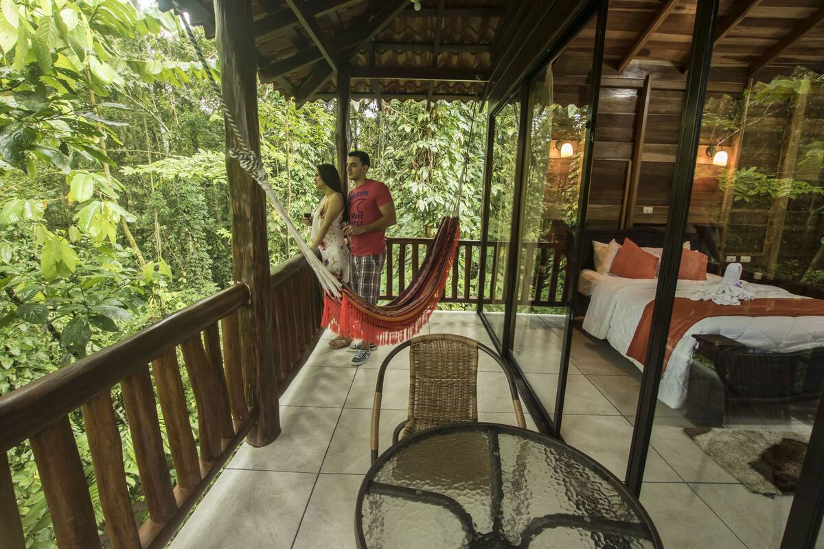 A wooden balcony with a glass table and chairs, overlooking a lush garden.