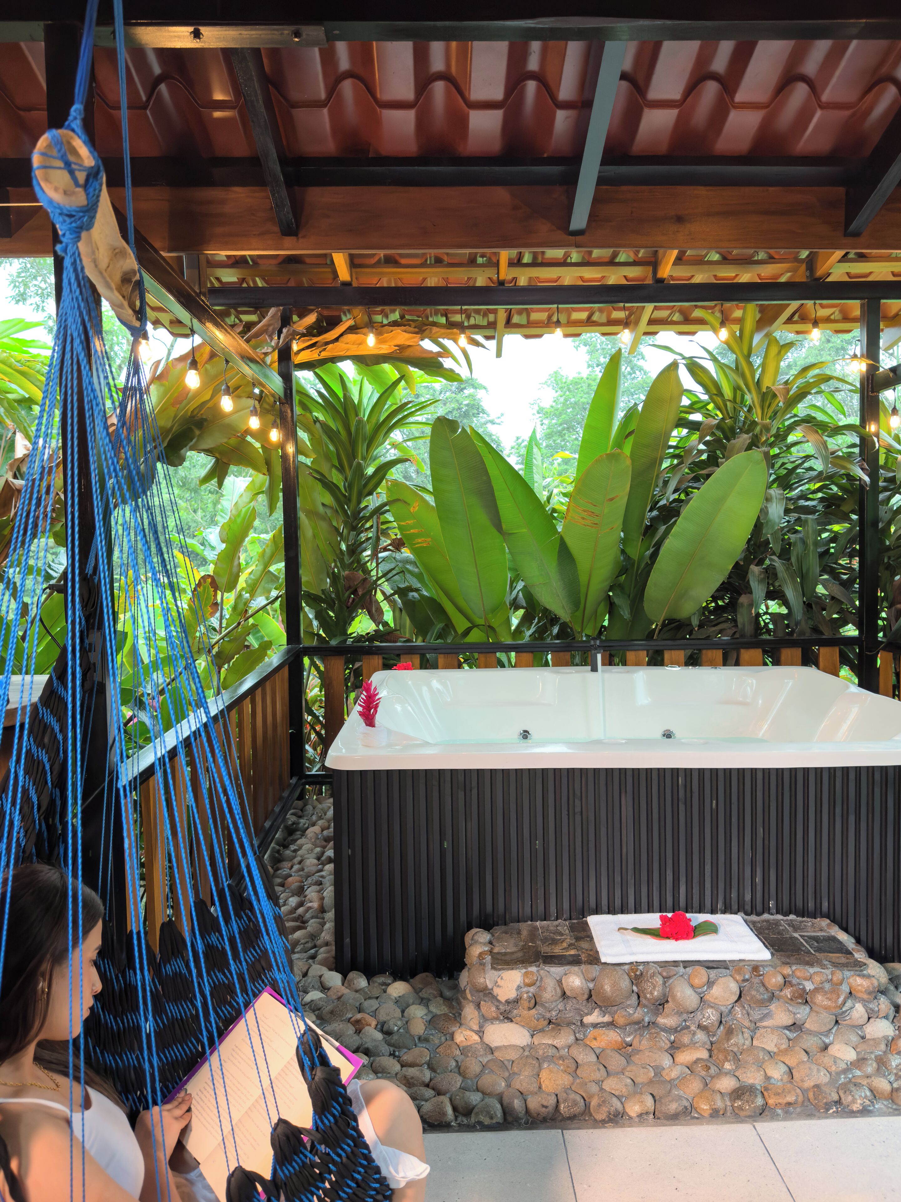 A hammock under a wooden pergola, a hot tub, and lush greenery.