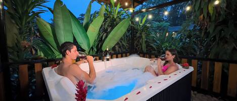 A couple relaxing in a hot tub surrounded by lush greenery and string lights.