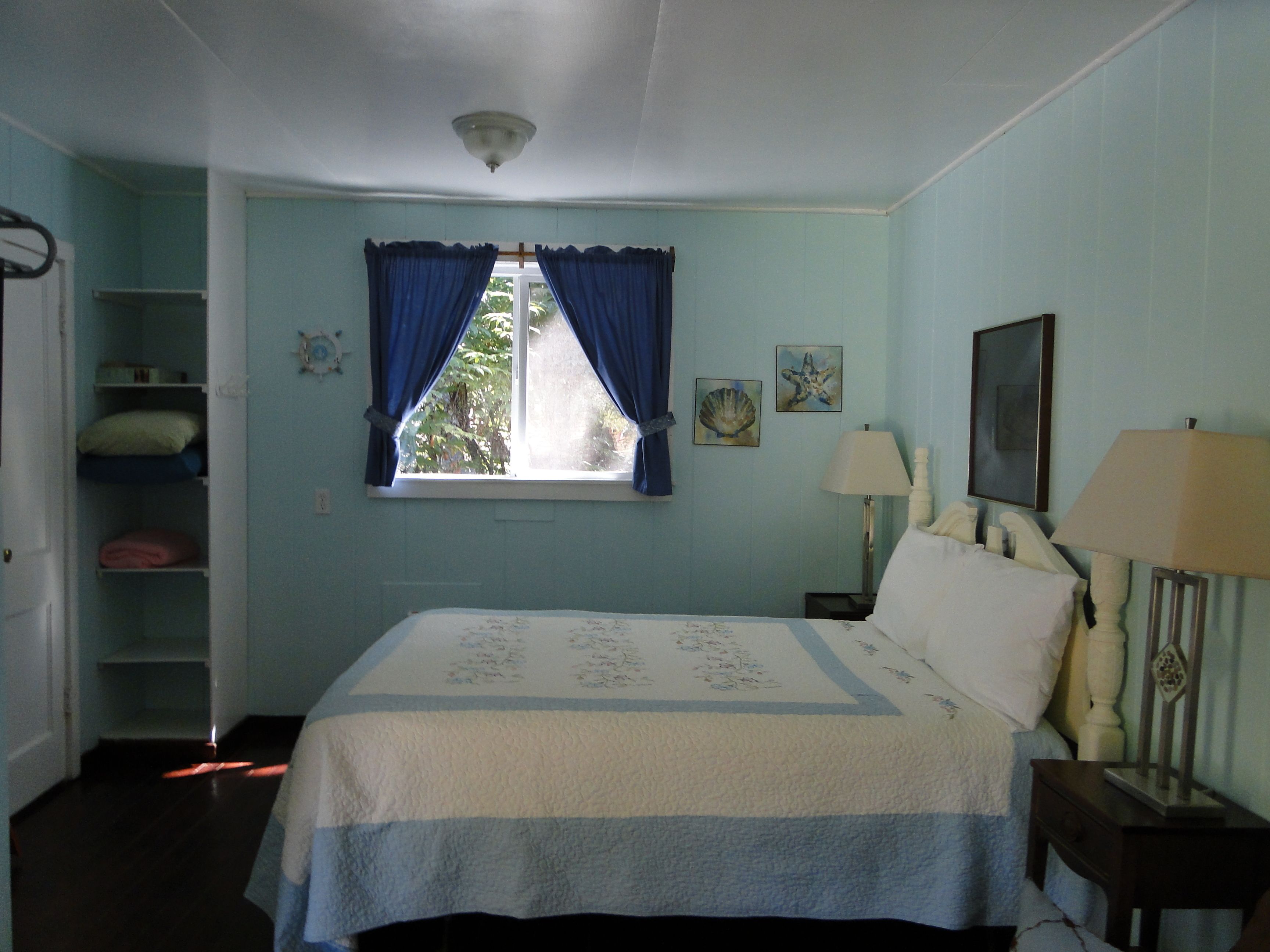 Une chambre à coucher avec un lit, deux lampes de chevet, une fenêtre avec des rideaux bleus, une étagère fixée au mur et un luminaire au plafond.