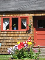 A wooden house with a red door and windows. There are potted plants on the windowsills.