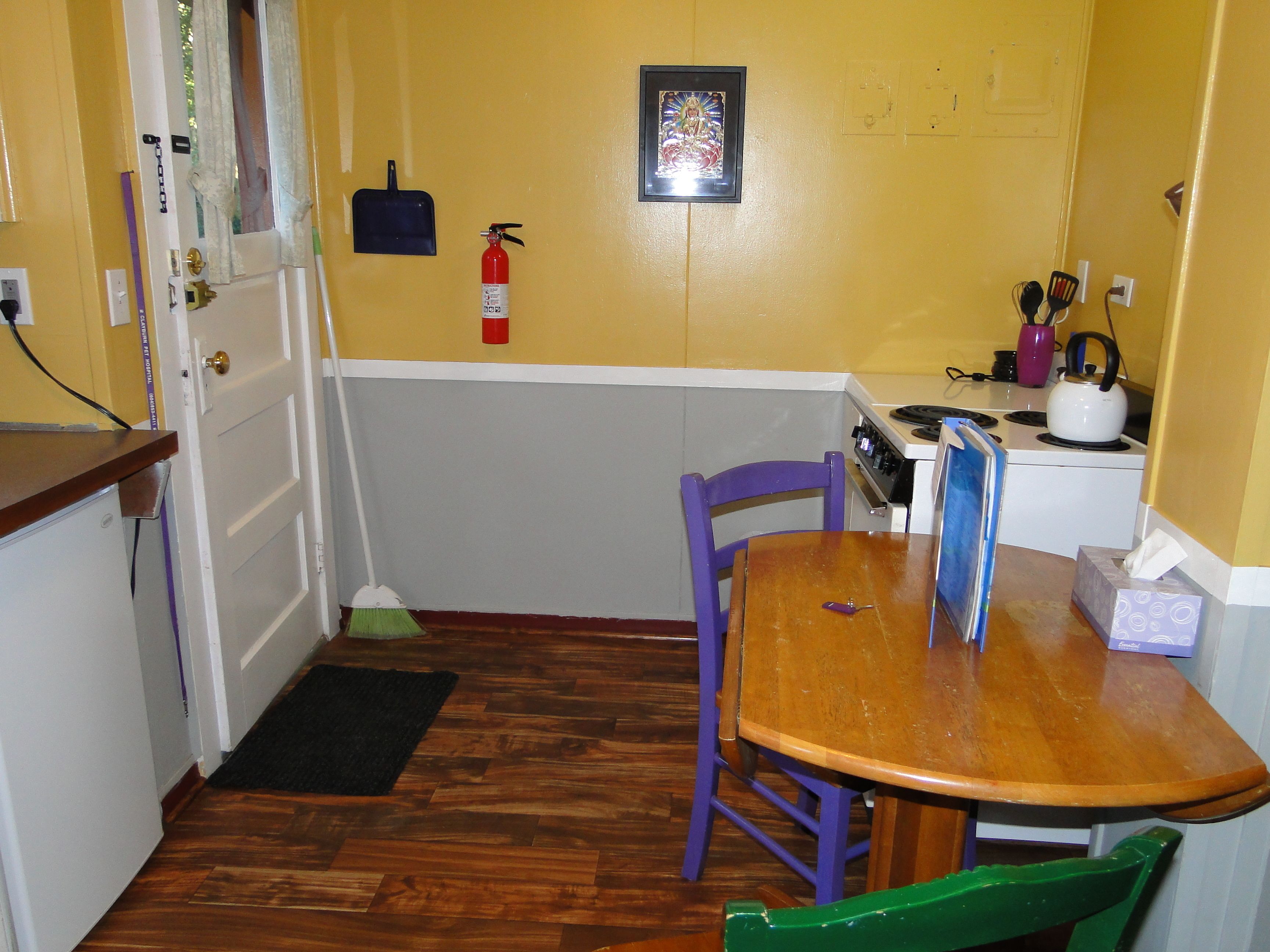 A small kitchen with a wooden table, purple chairs, a fire extinguisher, and a stove.
