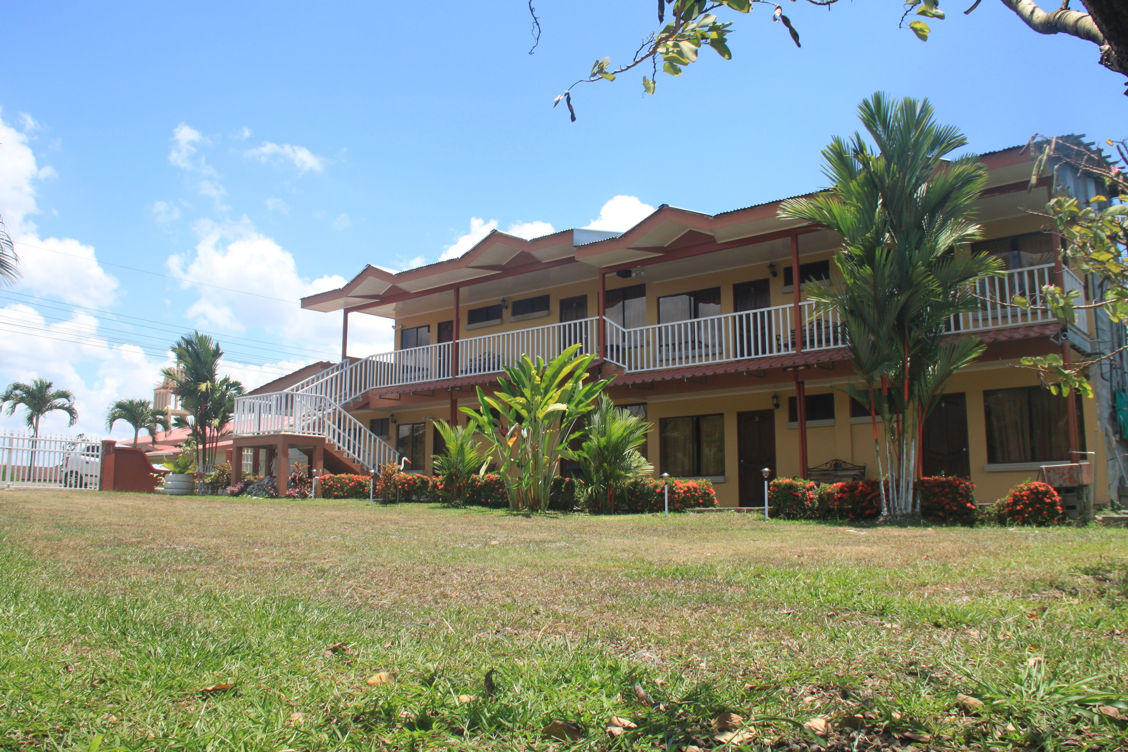 A multi-story hotel with a balcony on the second floor, surrounded by well-maintained lawns and tropical plants.