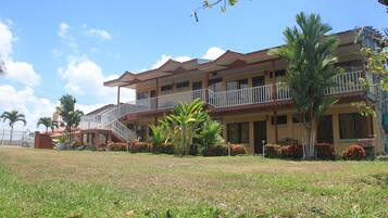A multi-story hotel with a balcony on the second floor, surrounded by well-maintained lawns and tropical plants.