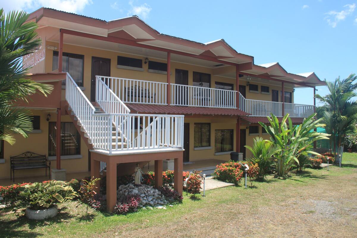 A two-story building with a balcony, a staircase, and a front garden with various plants and flowers.