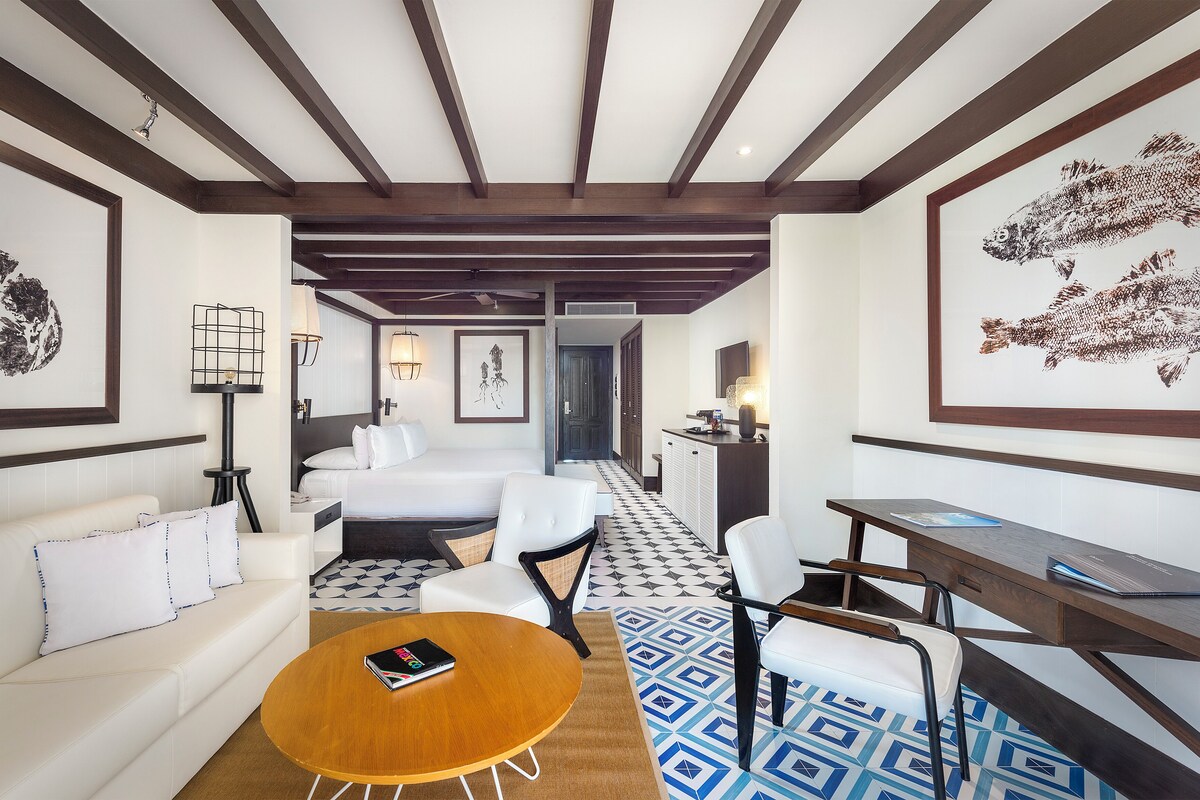 A hotel room with a bed, a sofa, a desk, and a coffee table. There is a patterned floor and a ceiling with wooden beams.