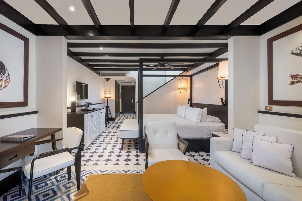 A modern hotel room with a sofa, a dining table, a desk, and a staircase leading to another room.