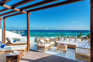 A covered outdoor seating area with lounge chairs and sofas, overlooking a beach and ocean.