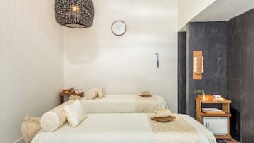 Une chambre d’hôtel avec deux lits, une horloge murale, une petite table d’appoint en bois et une étagère fixée au mur, sur laquelle se trouvent une plante et une horloge.