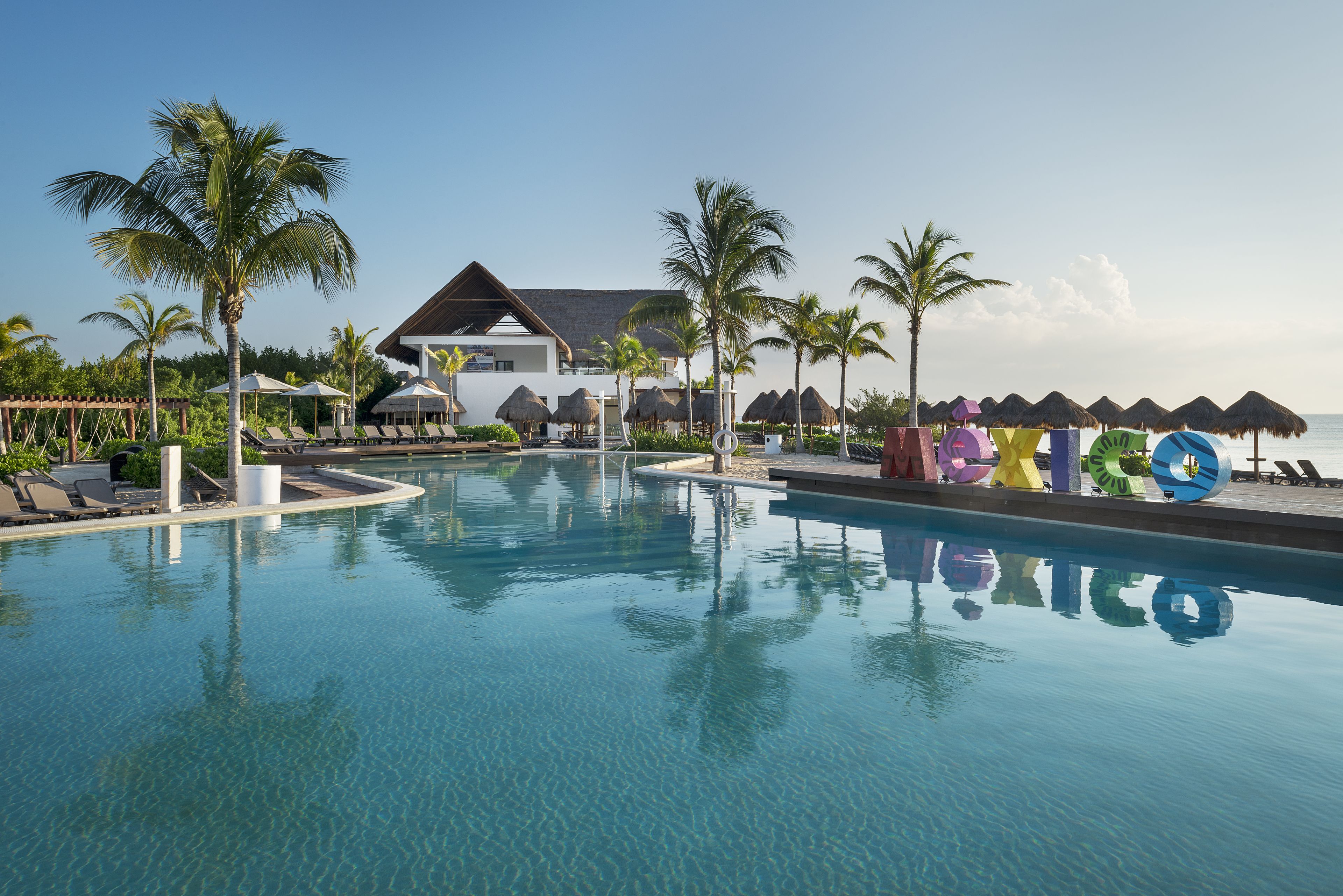 A resort pool with lounge chairs, palm trees, and a building with a thatched roof.