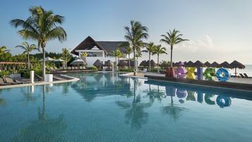 A resort pool with lounge chairs, palm trees, and a building with a thatched roof.