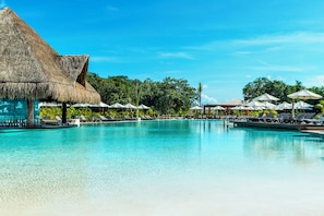 A clear blue pool with lounge chairs and umbrellas, surrounded by trees and a thatched-roof structure.