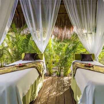A room with two beds, each with a pillow and a decorative item on the bed. The room has a wooden floor and is surrounded by greenery.