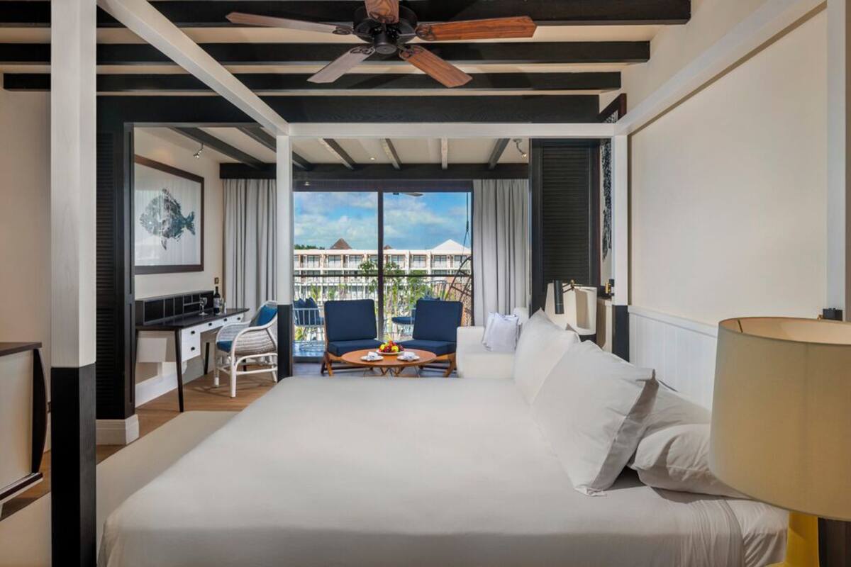 A hotel room with a bed, a desk, two chairs, a table with fruit, and a balcony with a view.