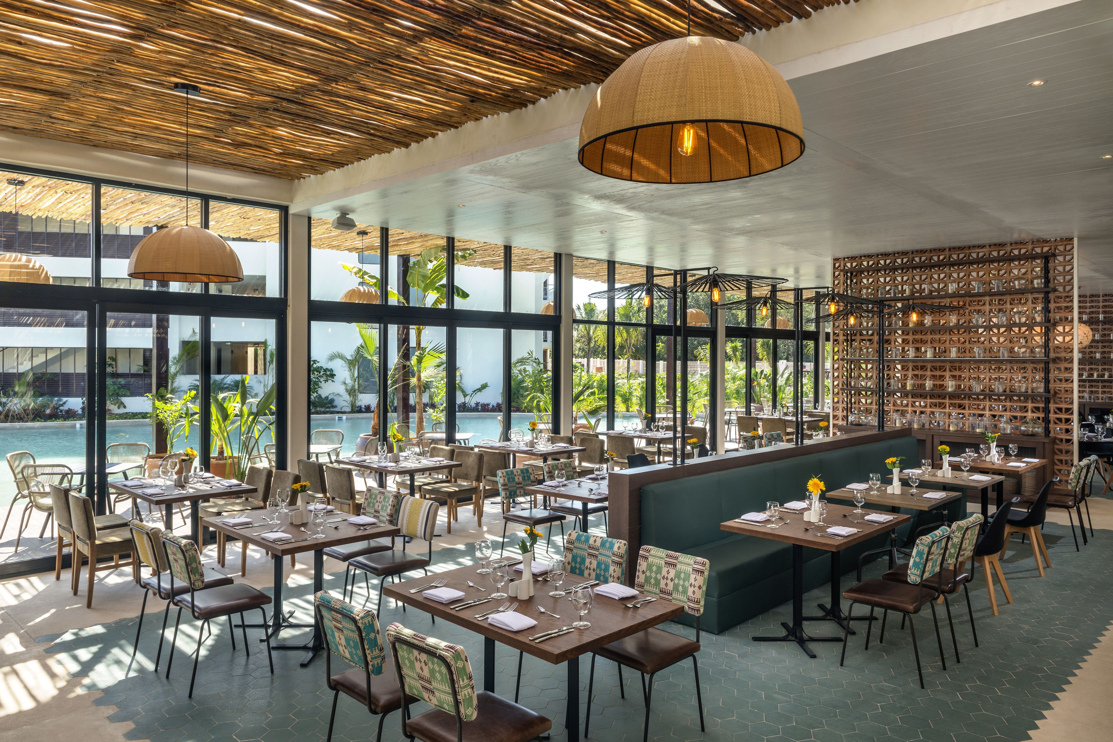 A restaurant with tables and chairs arranged for dining, large windows offering views of the outdoors, and a ceiling with a woven design.