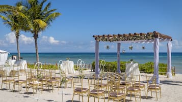 Een trouwdecoratie op het strand met een houten pergola, witte draperingen en gouden stoelen.