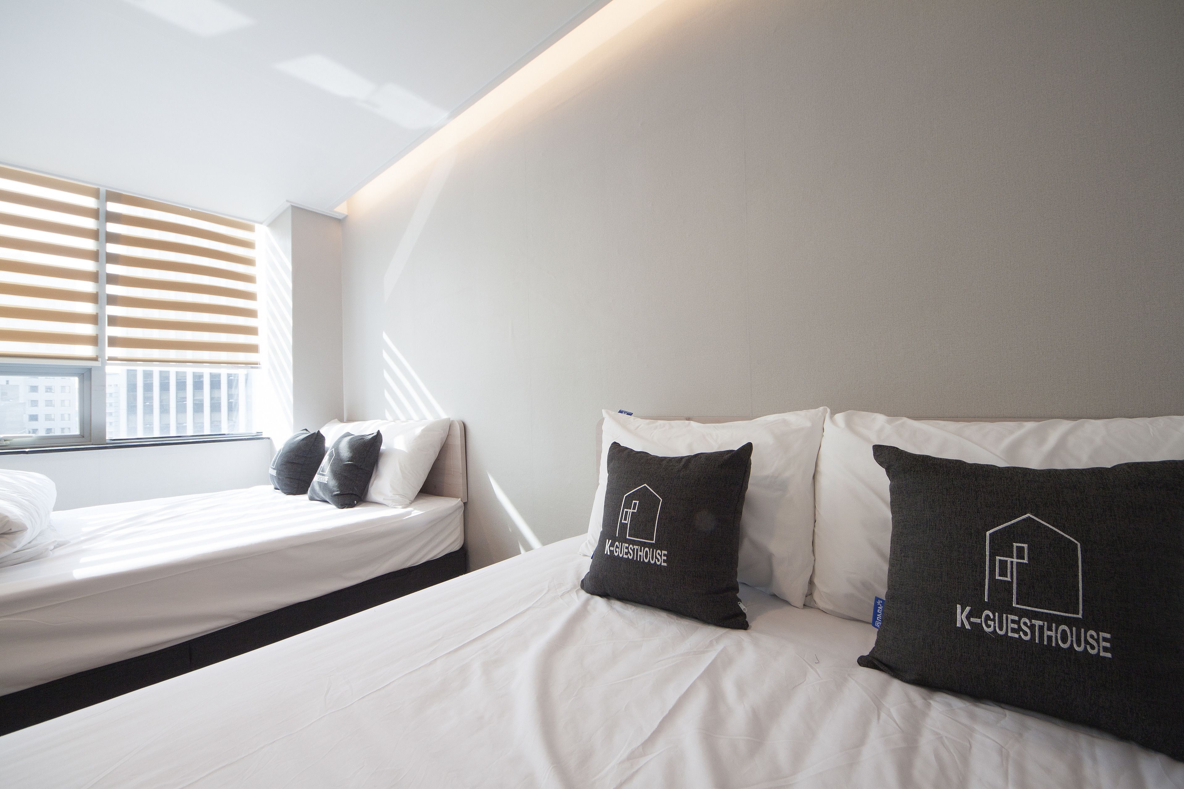 A modern hotel room with a bed, two pillows with 'K-GUESTHOUSE' branding, a window with blinds, and a city view.